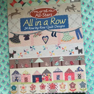 Könnte beinhalten: Ein Buch mit dem Titel "All in a Row" von Moda All-Stars, mit 24 Quilt-Designs Reihe für Reihe. Das Cover zeigt einen Patchwork-Quilt mit verschiedenen Designs, darunter Kleidung, Herzen, Hunde, Häuser und Blumen. Der Hintergrund ist ein türkis-rosa gemusterter Stoff.