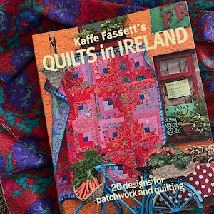Puede incluir: Un libro titulado "Kaffe Fassett's Quilts in Ireland" con un diseño de patchwork colorido en la portada. El libro está sobre una tela estampada con motivos florales rojos, morados y azules. También se ve una bicicleta con una cesta de zanahorias.