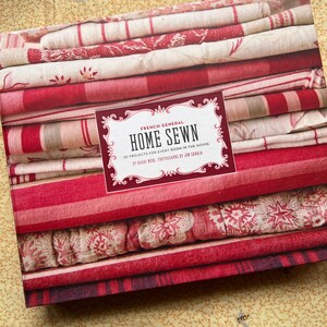 Può includere: Un libro intitolato "HOME SEWN" di French General, con una pila di tessuto rosso e bianco. La copertina del libro mostra il titolo in una cornice decorativa. I tessuti presentano vari motivi, tra cui righe e disegni floreali.