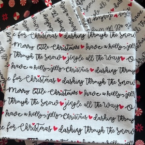 May include: A white fabric featuring repeating black cursive text that reads "dashing through the snow," "merry little Christmas," "have a holly jolly Christmas," and "jingle all the way," interspersed with small red heart icons throughout the pattern.