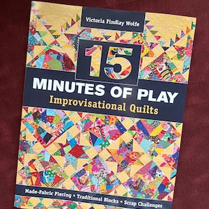 May include: A book titled "15 Minutes of Play Improvisational Quilts" by Victoria Findlay Wolfe. The cover features a colorful patchwork design with the number 15 in large, bold letters. The book promotes made-fabric piecing, traditional blocks, and scrap challenges.
