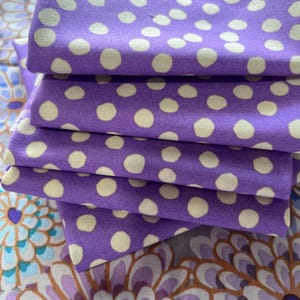 Può includere: Una pila di quadrati di tessuto piegati caratterizzati da uno sfondo viola con un motivo ripetitivo di piccoli pois color crema chiaro. Il tessuto ha una trama visibile ed è stratificato per mostrare più pezzi identici impilati insieme.