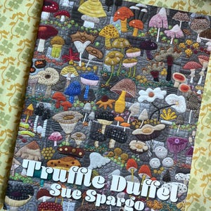 Può includere: Un'opera d'arte tessile raffigurante un dettagliato design di funghi in vari colori e trame. L'opera d'arte include il testo "Truffle Duffel Sue Spargo" in lettere bianche, mostrando una varietà di specie di funghi.