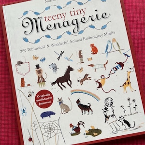 Puede incluir: Libro de Noriko Komurata, "teeny tiny Menagerie", con 380 motivos de bordado de animales. La portada muestra animales bordados, como un gato, pájaros y una jirafa. Texto "Originally published in Japanese!" sobre fondo rojo.