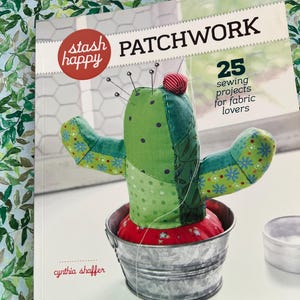 Op de afbeelding: Een boek getiteld "PATCHWORK" met een groen cactusvormig speldenkussen op de cover. Het boek heeft de woorden "stash happy" in een rode cirkel en "25 naaiprojecten voor stoffen liefhebbers". De cactus staat in een metalen emmer.