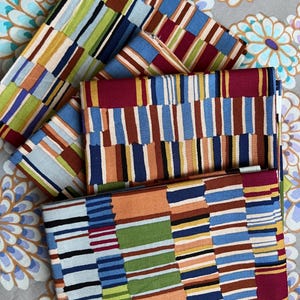May include: A stack of folded fabric pieces featuring a repeating pattern of vertical stripes in various widths and colors, including shades of blue, orange, green, red, yellow, and black, arranged in a textured, block-like composition.