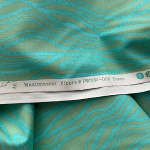 以下が含まれることがあります： ターコイズとグリーンの色合いで、波のような流れるような模様の生地のクローズアップ。生地は柔らかく、質感のある外観です。「Westminster Fibers # PWNW-060 Dunes」というテキストが白いラベルに印刷されています。