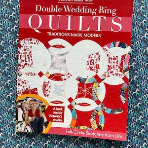 Può includere: Un libro rosso intitolato "Double Wedding Ring Quilts" di Victoria Findlay Wolfe. La copertina presenta un disegno di quilt con cerchi bianchi e dettagli rossi. Il libro include anche il testo "Traditions Made Modern" e "Full-Circle Sketches from Life."