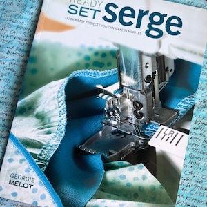 May include: A softcover book titled Ready Set Serge: Quick & Easy Projects You Can Make In Minutes by Georgie Melot. The cover features a close-up of a serger sewing machine stitching blue and teal polka-dot fabric against a cat-themed background.
