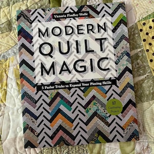Könnte beinhalten: Ein Buch mit dem Titel "Modern Quilt Magic" von Victoria Findlay Wolfe. Das Cover zeigt ein Zickzackmuster in verschiedenen Farben und den Titel in großen schwarzen Buchstaben. Das Buch verspricht 5 Tricks zur Erweiterung der Nähfähigkeiten und 17 fesselnde Projekte.