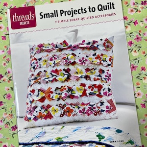 Op de afbeelding: Een boek getiteld "Small Projects to Quilt" van Threads Selects, met een kleurrijk, uit restjes gemaakt kussen met bloemaccenten. Het kussen is wit met lagen stof in rood, geel en oranje. De achtergrond heeft een bloemenpatroon.