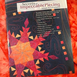May include: A softcover instructional book titled Secrets of Impeccable Piecing by Susan K. Cleveland. The cover features a dark blue background with a vibrant orange and red star quilt pattern, white text, and a list of quilting techniques.