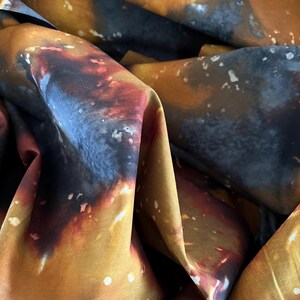 May include: Close-up of a fabric with a swirling, abstract design. The fabric features a blend of colors, including shades of brown, orange, and blue, with white speckles. The fabric is folded, creating shadows and depth.