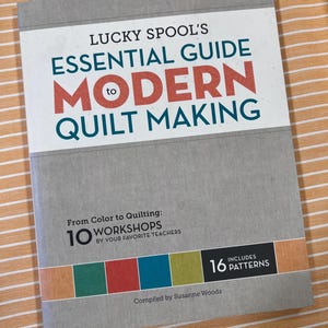 Op de afbeelding: Een boek getiteld "Lucky Spool's Essential Guide to Modern Quilt Making" met rode en blauwgroene tekst. De omslag is grijs met een witte banner. Het bevat 10 workshops en 16 patronen. Samengesteld door Susanne Woods.