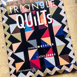 Può includere: Un libro intitolato "Free Range Triangle Quilts" con un design geometrico colorato sulla copertina. La copertina presenta uno sfondo nero con triangoli di vari colori, tra cui rosa, blu e arancione. Gli autori sono Gwen Marston e Cathy Jones.