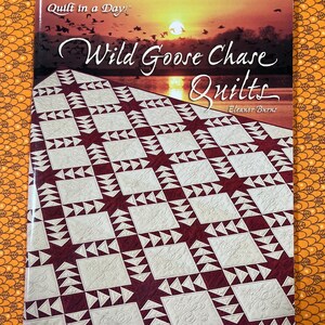 May include: A book titled "Wild Goose Chase Quilts" by Eleanor Burns. The cover features a sunset scene with geese in flight and a quilt pattern. The quilt design is a burgundy and cream color scheme.