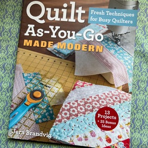 Peut inclure: Un livre intitulé "Quilt As-You-Go MADE MODERN" avec le sous-titre "Fresh Techniques for Busy Quilters". La couverture présente des blocs de courtepointe colorés, un cutter rotatif et le nom de l'auteur, Jera Brandvig. Le livre promet 13 projets et 25 idées bonus.