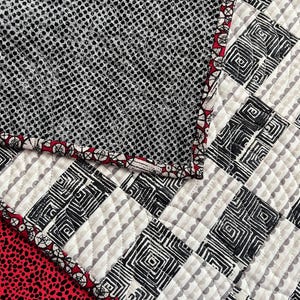 May include: A patchwork textile with a geometric design in black and white, a red border, and a red dotted fabric. The design incorporates squares and a dotted pattern, creating a visually interesting contrast.