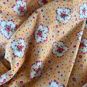 May include: Peach-coloured fabric with a floral pattern. The design features white circles with pink and red flowers, blue accents, and small black polka dots. The fabric is gathered, showing its texture and folds.
