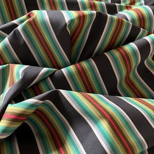 May include: Close-up of fabric with a repeating striped pattern. The stripes alternate between dark gray, white, light gray, green, yellow, pink, and brown. The fabric is folded, creating shadows and depth.