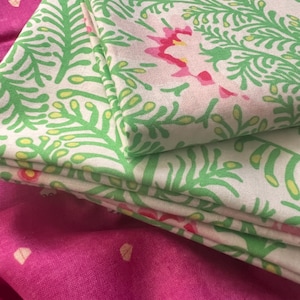 May include: A stack of folded white fabric featuring a repeating green fern-like leaf pattern with small pink and yellow floral accents, resting on top of a solid magenta fabric with a subtle, scattered gold geometric print.