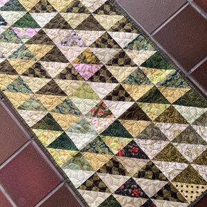 May include: A rectangular quilted table runner featuring a repeating triangular pattern in shades of olive green, mustard yellow, cream, and dark brown. The fabric pieces display various floral, polka dot, and textured prints with intricate stitching.