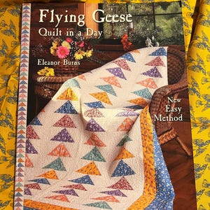 Op de afbeelding: Een boek getiteld "Flying Geese Quilt in a Day" van Eleanor Burns. De omslag toont een quilt met kleurrijke driehoekige patronen. De quilt is over een rieten stoel gedrapeerd. De achtergrond is een gele stof met een vogelpatroon.