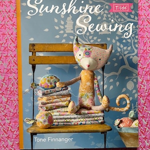 Puede incluir: Un libro titulado "Sunshine Sewing" de Tone Finnanger, con una muñeca de gato caprichosa sentada en un banco de madera. El gato está hecho de tela estampada, con paquetes de tela y un pequeño cubo cerca.