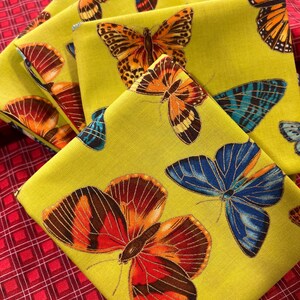 May include: A stack of square fabric napkins in a bright lime green colour featuring a repeating pattern of butterflies in shades of orange, red, and blue. The butterflies have detailed wing markings and are scattered across the fabric surface.