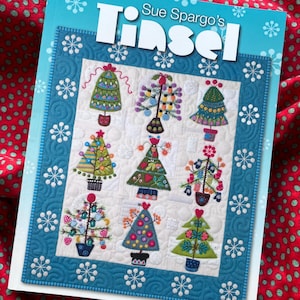 Peut inclure: Un livre à couverture souple intitulé Sue Spargo's Tinsel présentant un motif de courtepointe sur la couverture. La courtepointe affiche neuf motifs d'arbres de Noël brodés de différentes couleurs et formes sur un fond blanc avec une bordure bleue.