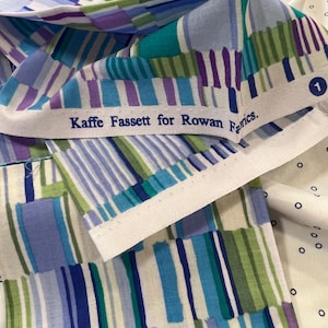 May include: A patterned fabric featuring vertical stripes in blue, green, and purple. The fabric includes the text "Kaffe Fassett for Rowan Fabrics" on a white border. The fabric is folded and layered.
