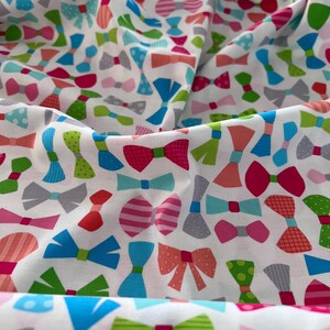 May include: White fabric featuring a repeating pattern of colourful bow ties in shades of pink, blue, green, orange, and grey. The bow ties have various designs, including polka dots and stripes. The fabric is folded, creating shadows and depth.