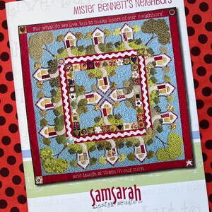 May include: A colorful quilt with a house and tree design, titled "Mister Bennett's Neighbors." The quilt features houses with red roofs and green trees on a blue background, framed by a red border. The text "Samsarah Design Studio" is visible.