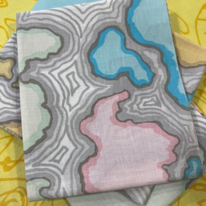 May include: Folded fabric napkins with a grey, pink, and blue abstract pattern. The napkins are stacked, with a yellow floral patterned fabric in the background. The napkins have a topographic map design, suitable for home decor.