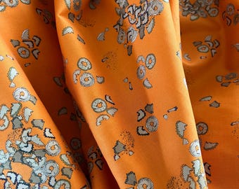 May include: Close-up of orange fabric with a repeating pattern of gray and white floral and abstract shapes. The fabric is folded, creating shadows and highlighting the texture and sheen of the material. The design is a mix of organic and geometric elements.