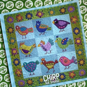 Peut inclure: Une courtepointe colorée avec neuf oiseaux brodés de différents motifs et couleurs, sur un fond bleu clair. La courtepointe est bordée de motifs floraux et de feuilles, avec le mot "CHIRP" et le nom "SUE SPARGO" affichés.
