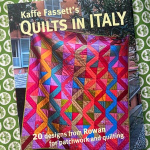 May include: A book titled "Kaffe Fassett's Quilts in Italy" featuring a colorful quilt design. The quilt has a geometric pattern with red, pink, blue, and orange hues. The book cover also includes text: "20 designs from Rowan for patchwork and quilting."