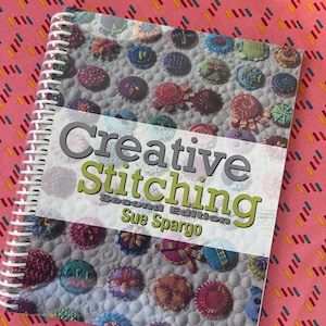 May include: A spiral-bound instructional book titled Creative Stitching Second Edition by Sue Spargo. The cover features a collage of small, colourful, circular embroidered fabric pieces arranged on a textured grey background with white stitching.