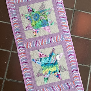 May include: A rectangular quilted table runner featuring three star-shaped blocks with vibrant floral patterns in shades of blue, green, and pink. The stars are set against a light beige background with quilted leaf motifs and a wavy striped border.