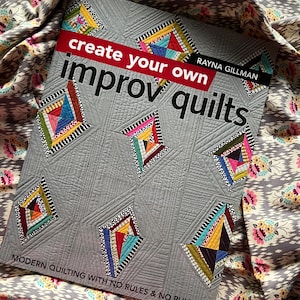 Könnte beinhalten: Ein graues Buch mit dem Titel "create your own improv quilts" mit farbenfrohen geometrischen Quilt-Designs. Das Buchcover hat ein rotes Banner mit weißem Text und ist von gemustertem Stoff umgeben.