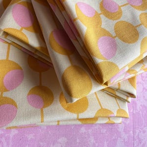 May include: Folded fabric featuring a repeating pattern of yellow and pink circles on an off-white backdrop. The fabric is stacked on a pink surface, creating a layered effect. The design has a retro aesthetic.