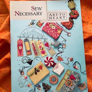 May include: A light blue craft book titled Sew Necessary by Art-to-Heart featuring illustrations of various fabric projects including small pouches, hanging ornaments, and decorative items with floral, heart, and candy motifs scattered across the cover.