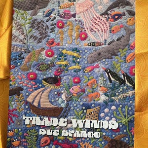 Pode incluir: Um edredom colorido com tema subaquático. O edredom apresenta várias criaturas marinhas, incluindo peixes, cavalos-marinhos, águas-vivas e um veleiro. O texto "TRADE WINDS SUE SPARGO" é exibido no edredom.