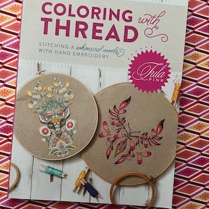 Könnte beinhalten: Ein Buch mit dem Titel "COLORING with THREAD" über Handstickerei. Das Cover zeigt zwei Stickrahmen mit floralen Designs und das Logo "Tula Pink". Garnrollen und Stickrahmen sind ebenfalls sichtbar.