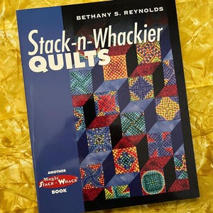 Puede incluir: Un libro titulado "Stack-n-Whackier Quilts" de Bethany S. Reynolds. La portada presenta un patrón de patchwork multicolor, con cuadrados rojos, azules, verdes y amarillos. El libro está etiquetado como "Another Magic Stack 'n Whack Book."