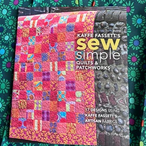 Könnte beinhalten: Ein Buch mit dem Titel Kaffe Fassett's Sew Simple Quilts & Patchworks mit einem rosa Patchwork-Quilt auf dem Cover. Das Cover zeigt den Text 17 designs using Kaffe Fassett's artisan fabrics und eine angrenzende Steinmauer.