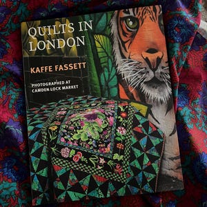 Könnte beinhalten: Ein Buch mit dem Titel "Quilts in London" von Kaffe Fassett, fotografiert am Camden Lock Market. Das Cover zeigt ein Tigergesicht und ein farbenfrohes Quilt-Design. Das Buch liegt auf einem Stoff mit roten, blauen und grünen Blumenmustern.