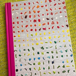 May include: A hardcover book with a bright pink spine and a light gray cover featuring a grid of small, raised, multi-colored shapes in a gradient pattern ranging from red and orange at the top to yellow, green, and blue at the bottom.