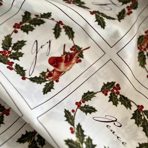 Può includere: Tessuto bianco con un motivo a griglia ripetuto di ghirlande di agrifoglio, bacche rosse e uccelli rossi. Ogni quadrato contiene una ghirlanda che circonda la parola Joy o Peace in corsivo nero. Il design include linee scure di separazione.