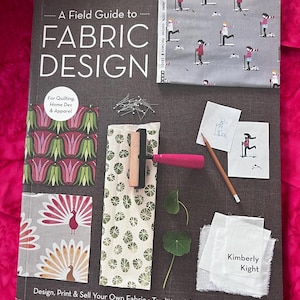 Könnte beinhalten: Ein Buch mit dem Titel A Field Guide to Fabric Design von Kimberly Kight. Das Cover zeigt Stoffmuster mit Blumen-, Pfauen- und Skifahrermotiven, eine Andruckwalze, Stecknadeln, einen Bleistift und zwei kleine Skizzen auf dunkelgrauem Grund.